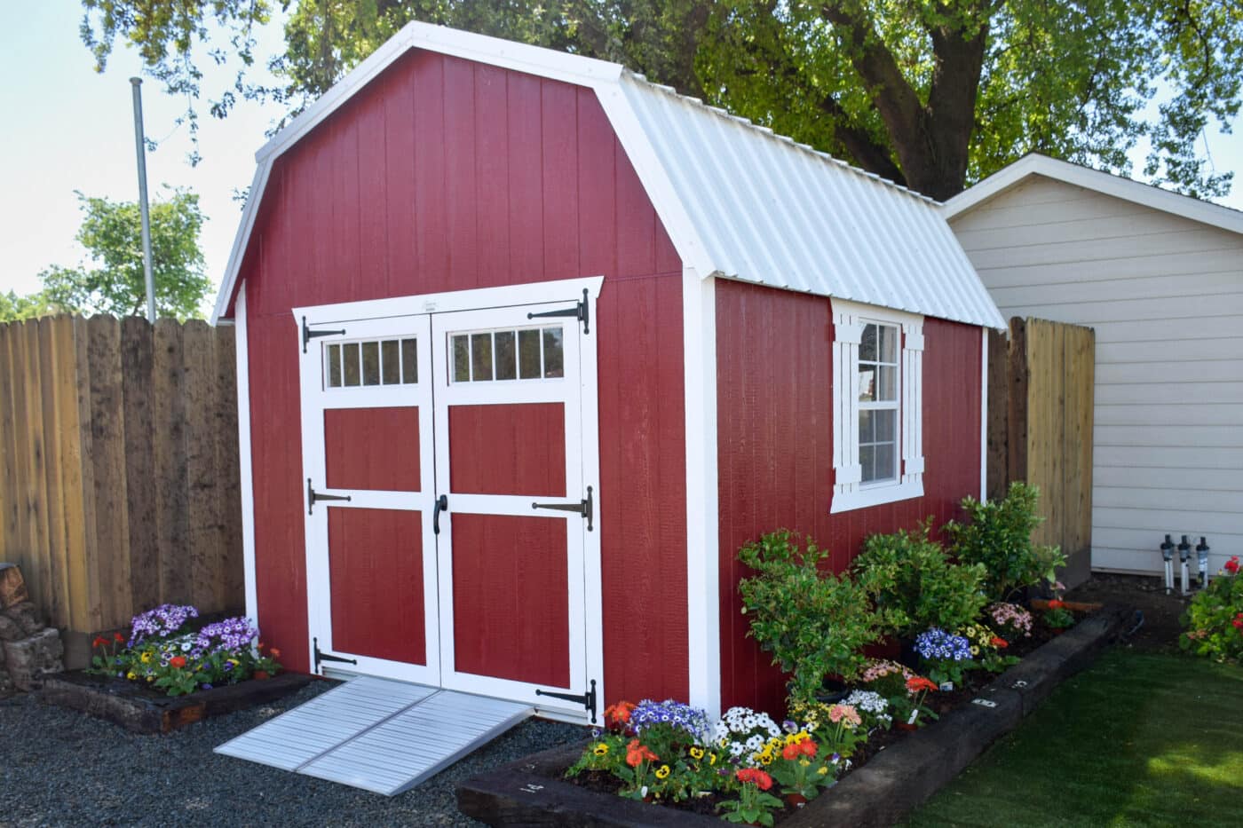 red and white trimmed barn elite shed with white metal roof and ramp surrounded by garden with purpole white yellow red blue and green flowers/plants