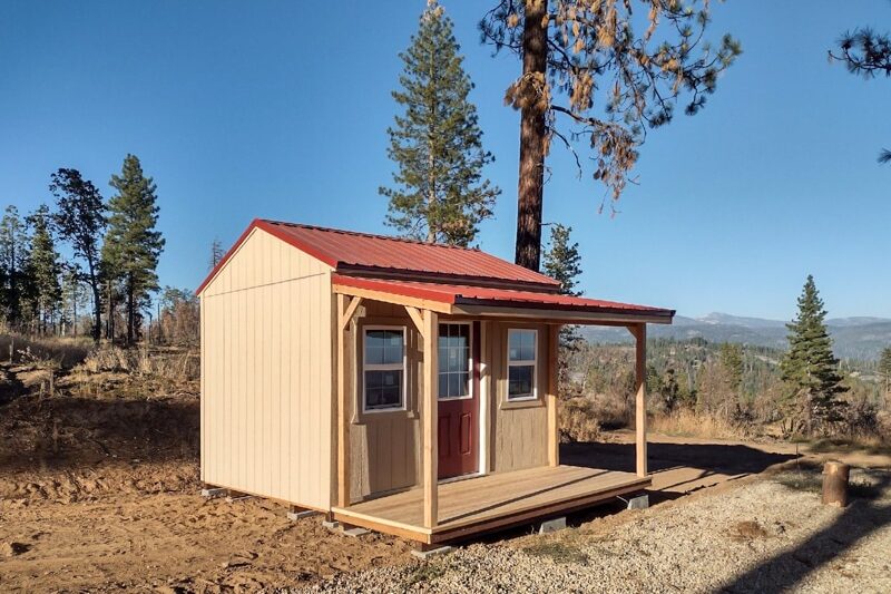 tan cottage shed with red metal roofing and overhang over front porch
