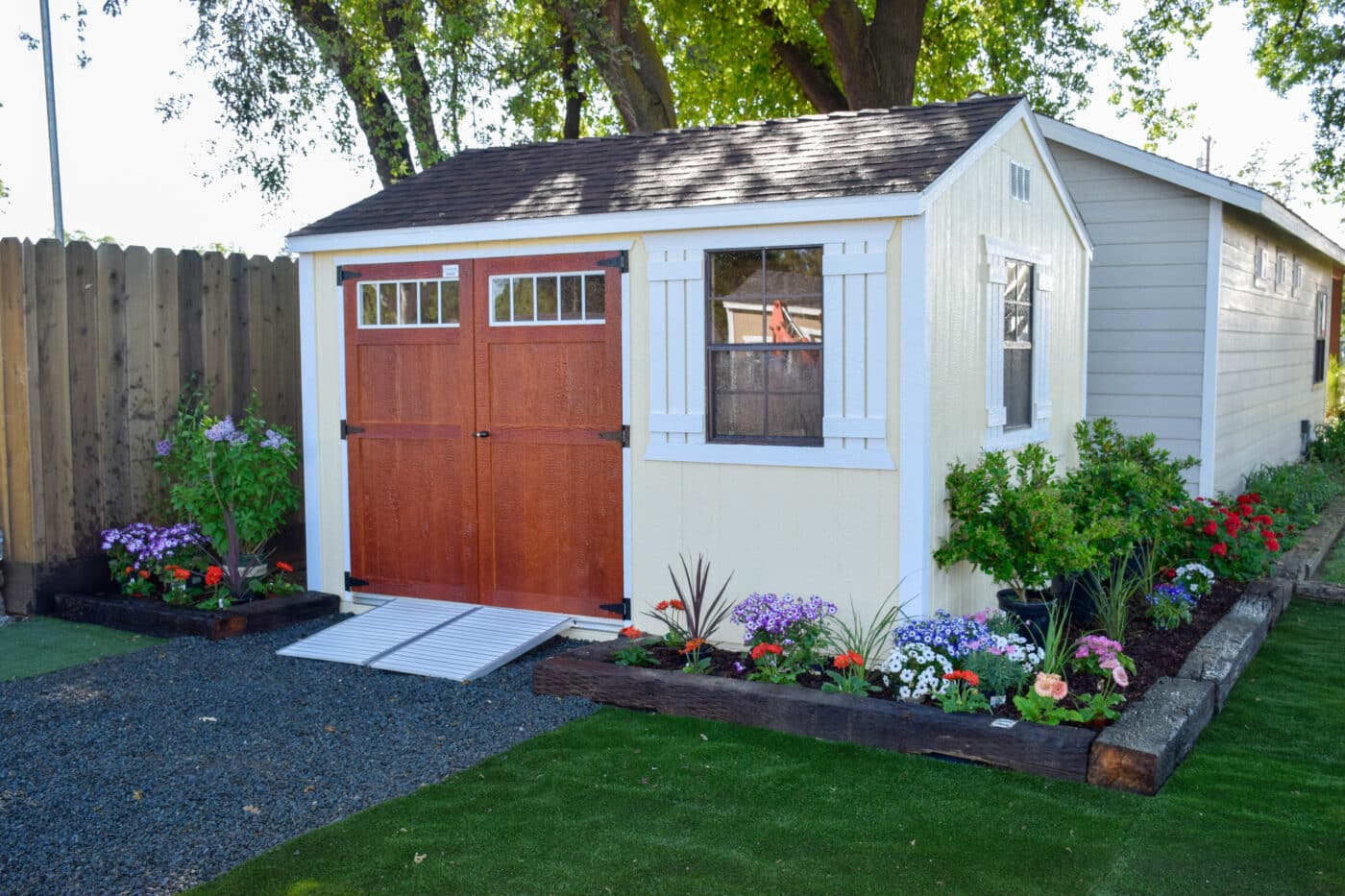 tan deluxe shed with reddish brown double door with ramp leading up to it surronded by garden bed